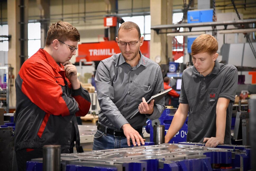 The picture shows the apprentice trainer explaining a punching and forming tool to two apprentices in toolmaking technology in a production environment. He points to specific parts of the tool and explains the functions and purpose of each component. The apprentices listen attentively and ask questions as they learn. The scene gives the impression of knowledge sharing and practical training in a professional working environment.