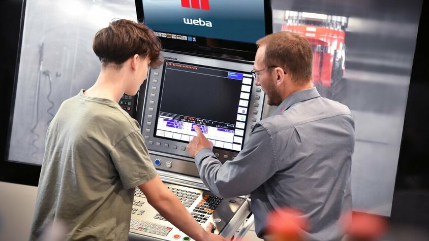 Ein Ausbilder erklärt einem Lehrling in der Zerspanungstechnik die Bedienfunktionen an der Steuerung eines Bearbeitungszentrums. In der Lehre in Steyr bei weba Werkzeugbau liegt der Fokus auf praxisnaher Ausbildung direkt an Maschinen.