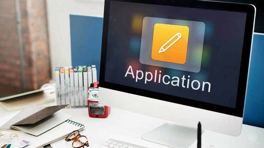  Desk with an Apple monitor, keyboard, and office supplies. On the screen, the word "Application" is displayed, symbolizing job openings in Steyr at weba Werkzeugbau.