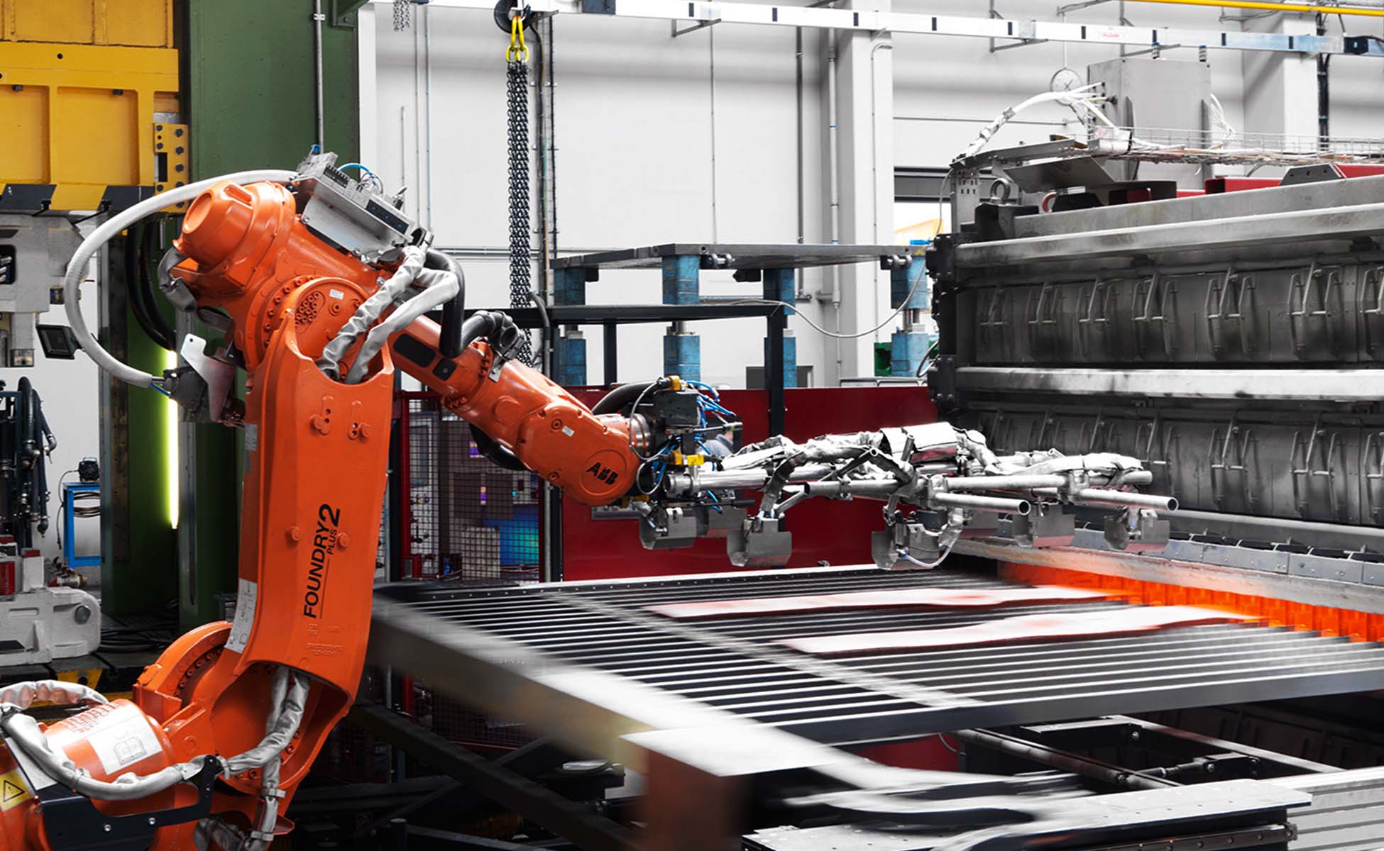  Ein Robotertransferarm greift zwei rotglühende Metallplatinen aus dem Inneren eines Kammerofens, um sie in ein Warmumformwerkzeug für die Herstellung von Strukturbauteilen zu transferieren. Die leuchtend roten Platinen stehen im Kontrast zum metallischen Glanz des Roboterarms und der dunkleren Umgebung des Industrieofens. Diese Szene veranschaulicht die Präzision und Automatisierung in modernen Fertigungsprozessen, insbesondere in der Herstellung hochfester Metallkomponenten für den Einsatz in anspruchsvollen Anwendungen.